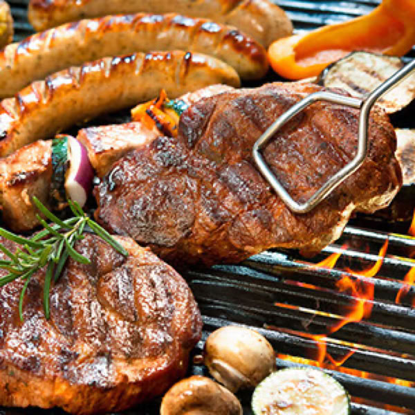 Gegrilltes Fleisch und W&uuml;rstchen auf einem Grill mit Flammen, umgeben von Gem&uuml;se wie Zucchini und Pilzen.
