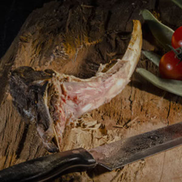 Ein Knochen mit etwas Fleisch auf einem Holzbrett, daneben ein Messer und zwei Tomaten mit Bl&auml;ttern.