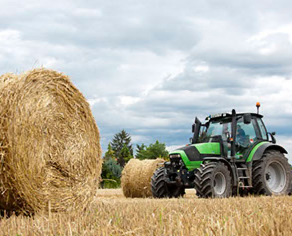 Ein gr&uuml;ner Traktor auf einem Feld mit gro&szlig;en Strohballen unter bew&ouml;lktem Himmel.