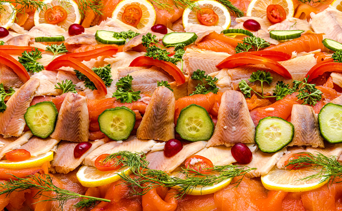 Verschiedene Fischsorten, Gurkenscheiben, Zitronenscheiben, rote Paprikastreifen, gekochte Eierh&auml;lften und Kr&auml;uter dekorativ auf einem Tablett arrangiert.
